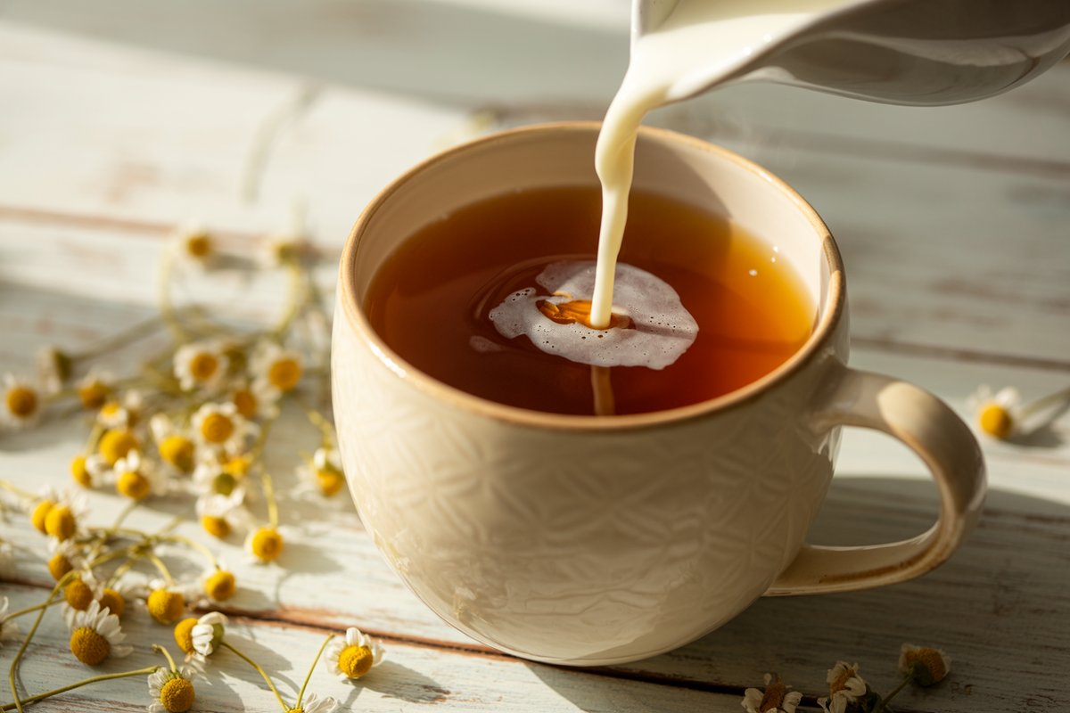 Hand pouring frothed milk into a mug of strong golden chamomile tea on a wooden counter