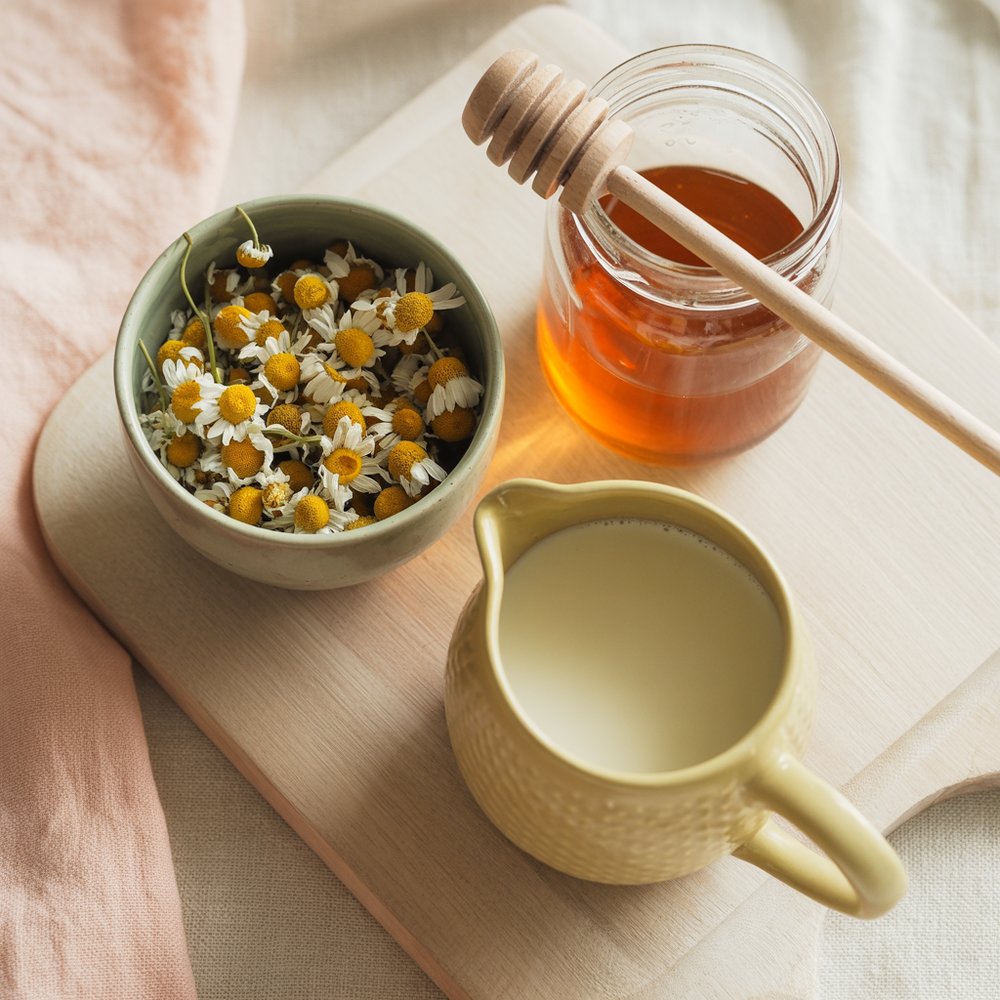 Overhead flat-lay of chamomile tea bags, loose chamomile buds, a honey jar, vanilla, and a small milk pitcher on a linen cloth