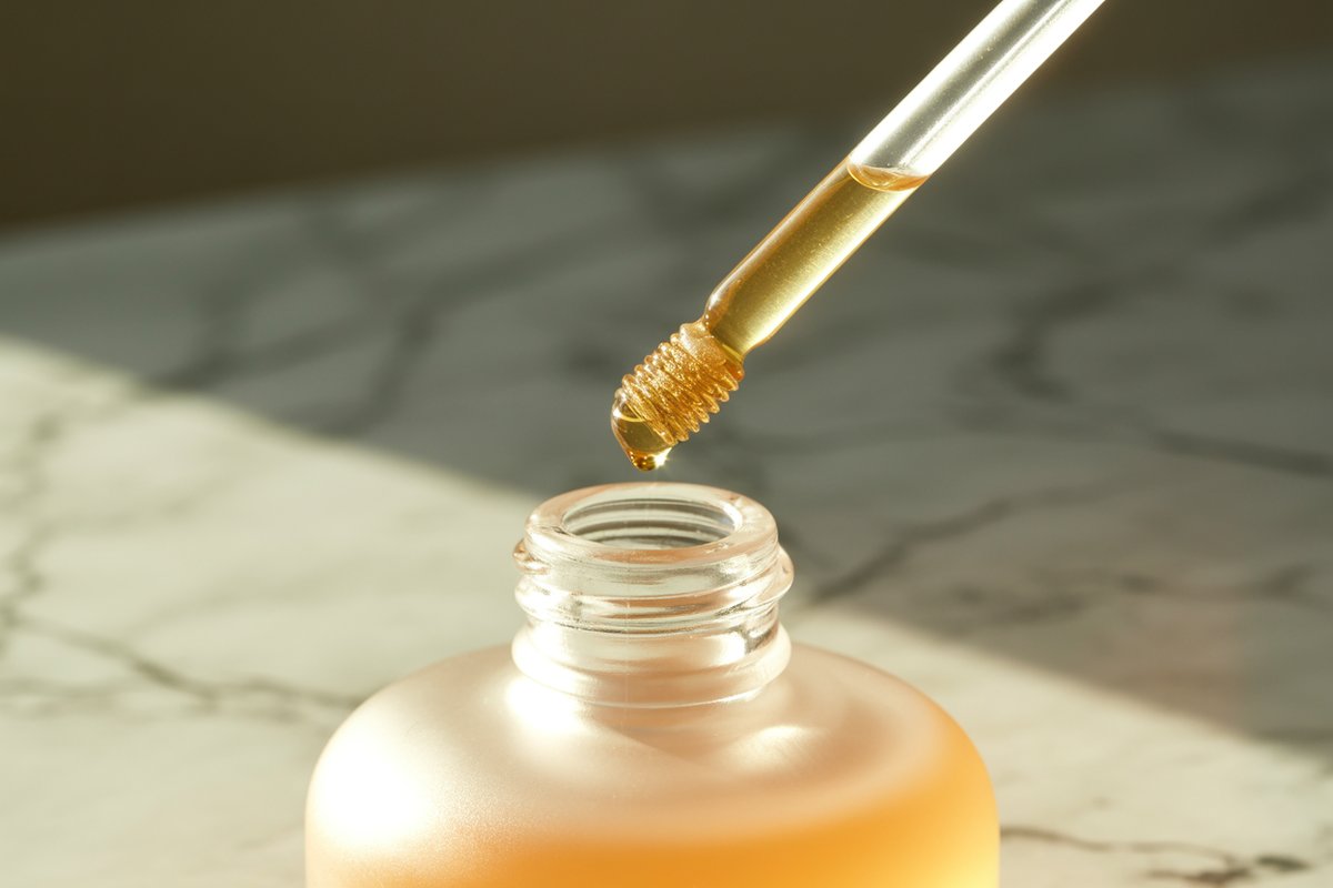 A doe-foot lip oil wand coated in glistening golden oil above an open glass bottle
